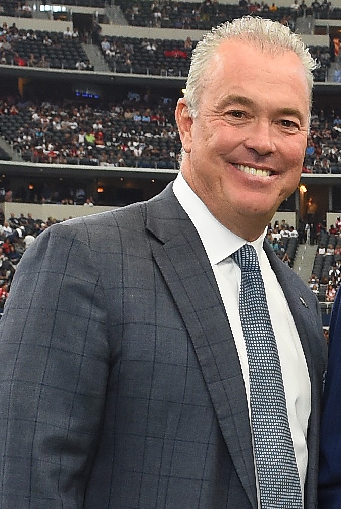 Man with grey hair and suit smiling in football stadium.
